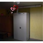 Pelēks apģērbu žāvēšanas skapis ar divām durvīm un ventilācijas izvadu, novietots telpā ar dzeltenām sienām.